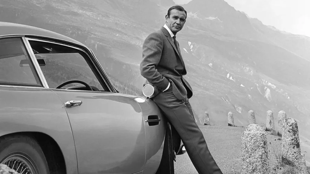 Feketefehér kép, ahogy Sean Connery James Bond-ként pózol az ikonikus Aston Martin mellett, a háttérben hegyek.
