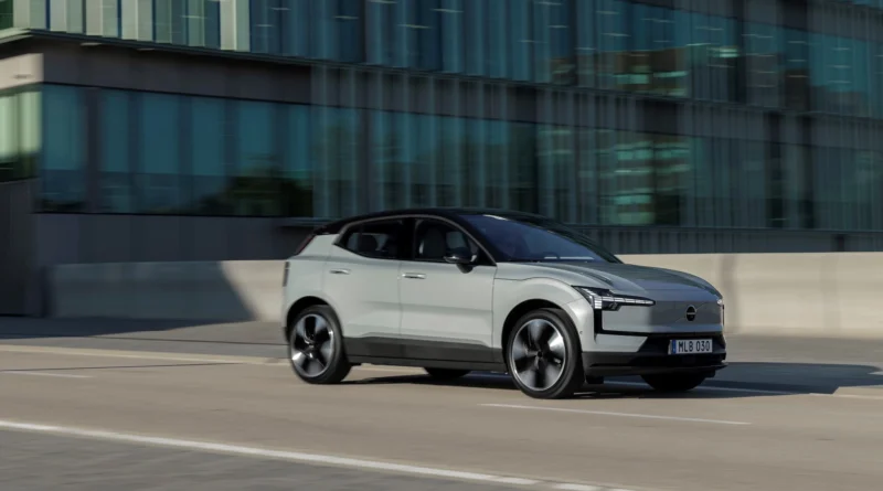 Szürke Volvo EX30 városban, egy üvegépület előtt halad az úton
